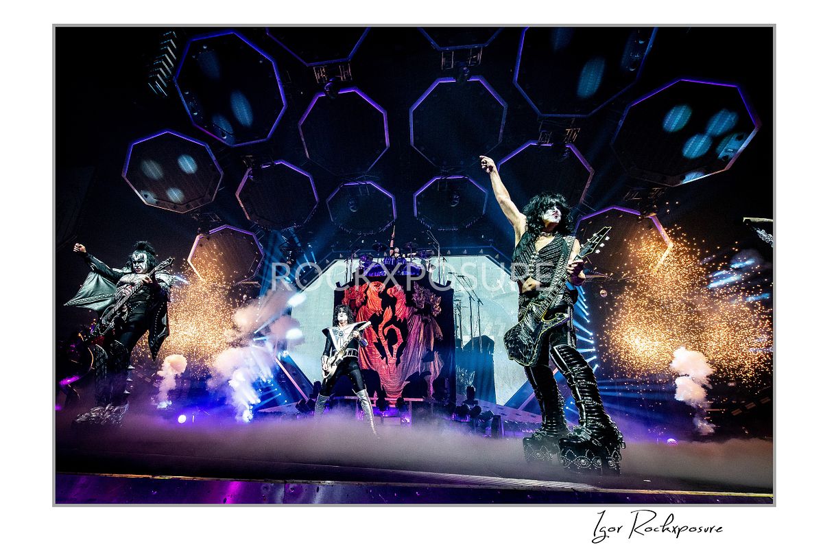 Horizontal color photograph of Gene Simmons, Paul Stanley, and Tommy Thayer performing onstage in full makeup and costumes with hexagonal lighting panels, red emblem backdrop, smoke, and pyrotechnic effects