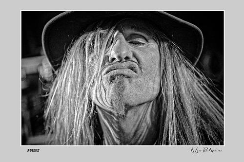 Horizontal black and white image of C.C. DeVille in an extreme close-up live portrait with long hair, hat and exaggerated expression