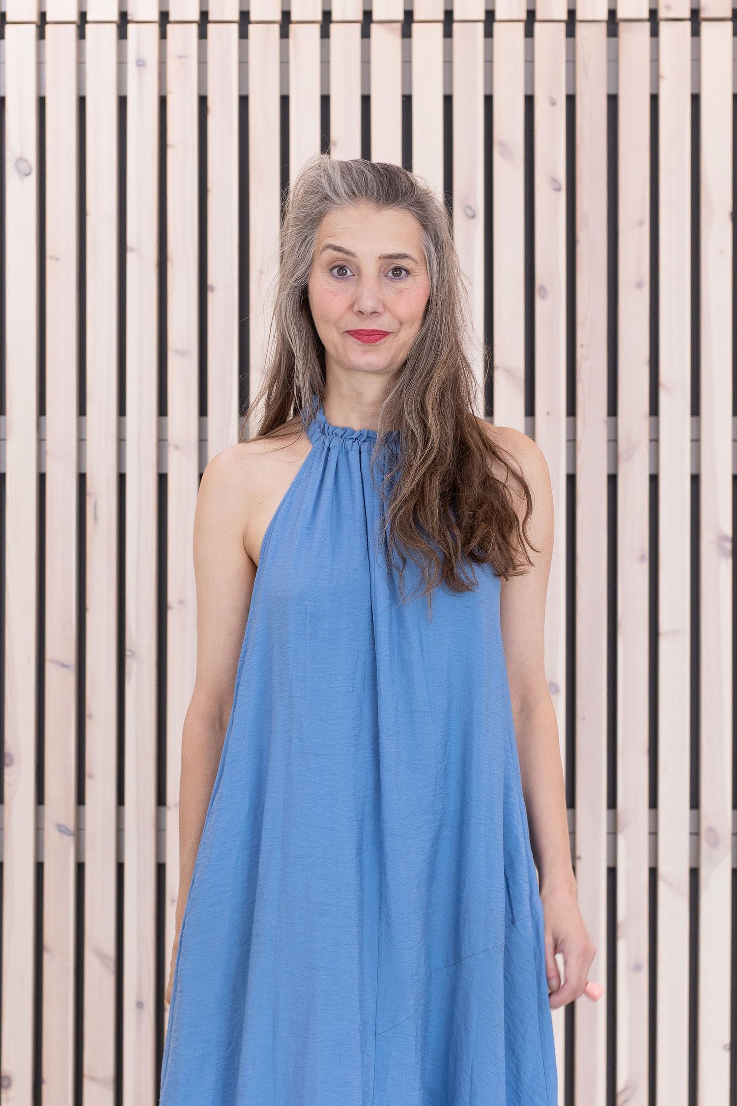 Natural light environmental portrait of a woman in a blue dress standing against a modern wooden slat wall, photographed in Stockholm by Mats Karlsson.