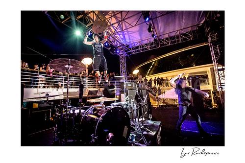 Horizontal color image of Zoltan Chaney leaping above the drum kit while holding a cymbal on stage during a live performance