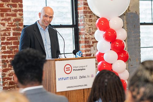 An Intuit executive speaks at a press conference for the IDEAS program, presented in partnership with the Urban League of Philadelphia, addressing a room of attendees in front of branded signage and balloons.