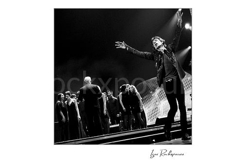 Square black and white image of Mick Jagger of The Rolling Stones performing beside a choir on stage during You Can’t Always Get What You Want
