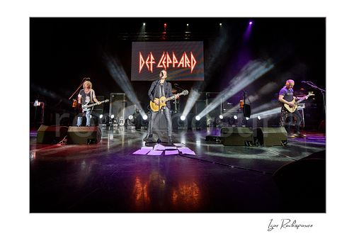 Horizontal color image of Rick Savage, Vivian Campbell, and Phil Collen of Def Leppard performing live on stage, with Savage at left on bass, Campbell at center on guitar, and Collen at right on guitar under bright concert lighting with the Def Leppard logo visible in the background