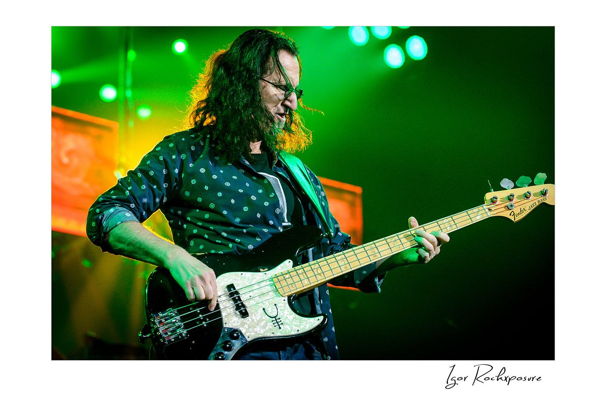 Horizontal concert photography of Geddy Lee playing bass guitar under green stage lighting