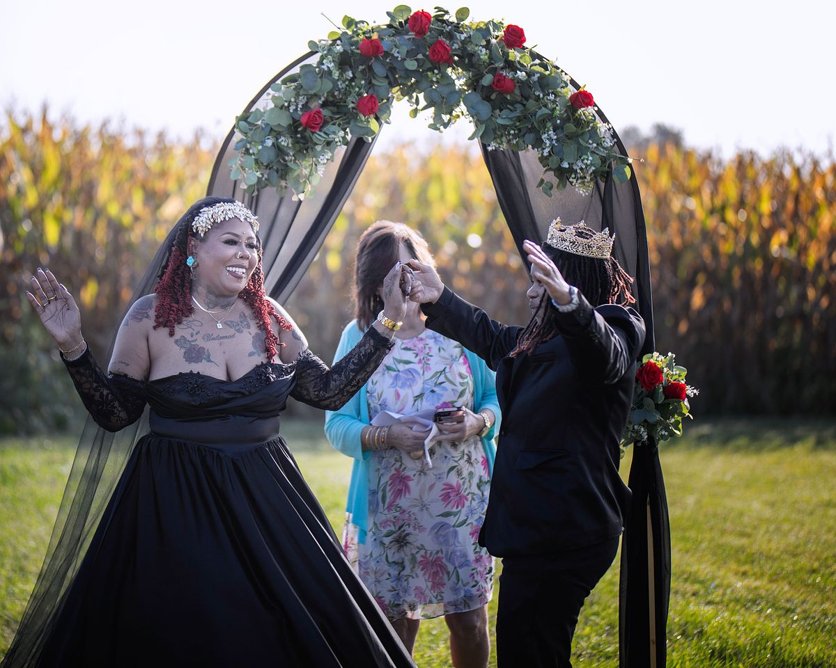 A true-to-color outdoor wedding ceremony featuring a couple in black attire celebrating under a floral arch in a sun-drenched field.