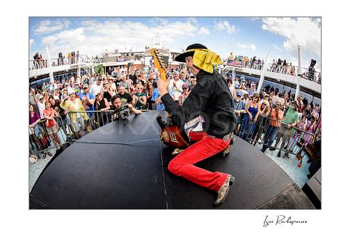 Horizontal color image of Randy Hansen kneeling on an outdoor ship deck stage with guitar and a large crowd behind him