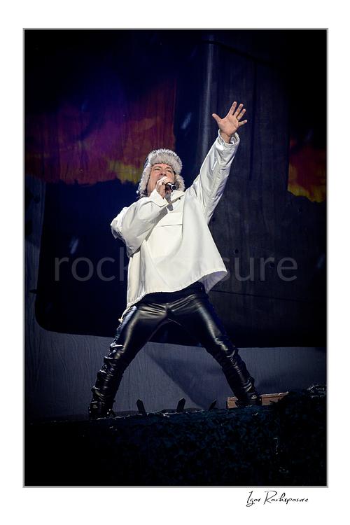 Vertical color image of Bruce Dickinson of Iron Maiden performing live on stage in a white outfit, singing into a microphone with one hand raised