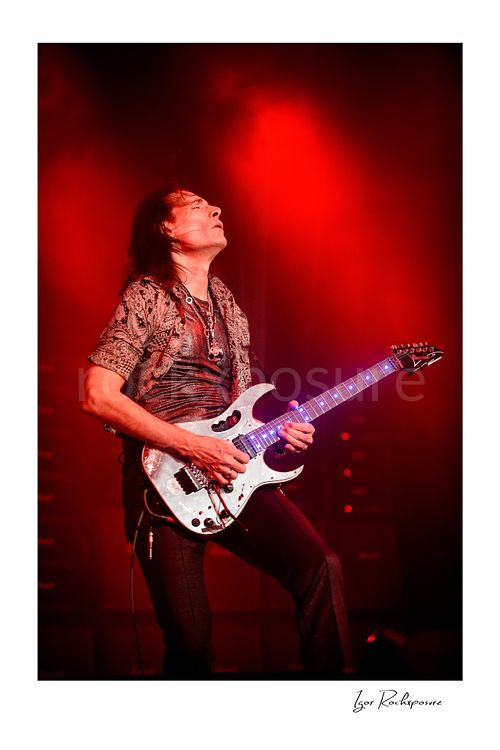 Vertical color image of Steve Vai playing guitar under vivid red stage lighting with glowing fret markers and head tilted back