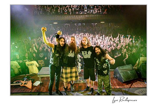 Wide shot of the four members of Black Label Society standing together on stage posing for the camera, with a large crowd behind them under green stage lighting.