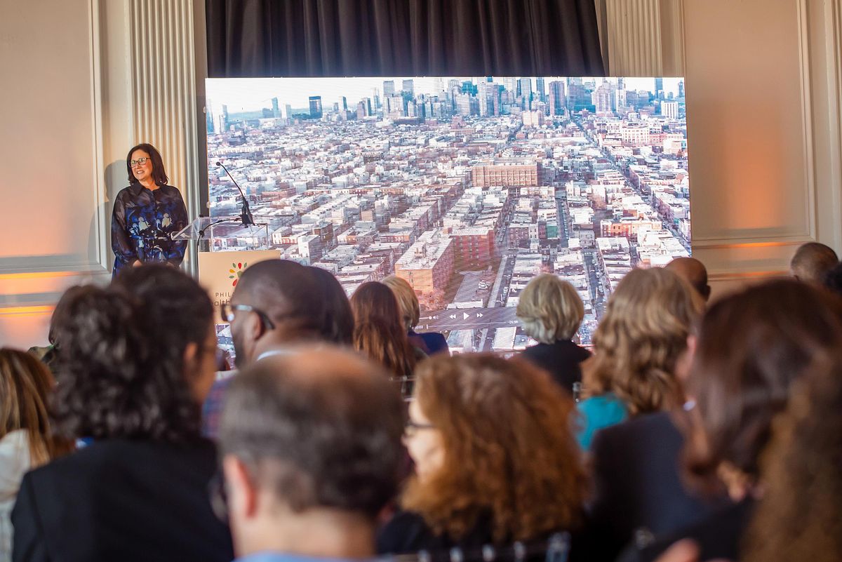 Corporate event photography capturing a keynote speaker addressing a professional audience at The Downtown Club in Philadelphia, framed by a digital city skyline backdrop, highlighting leadership, narrative presence, and a sense of place.