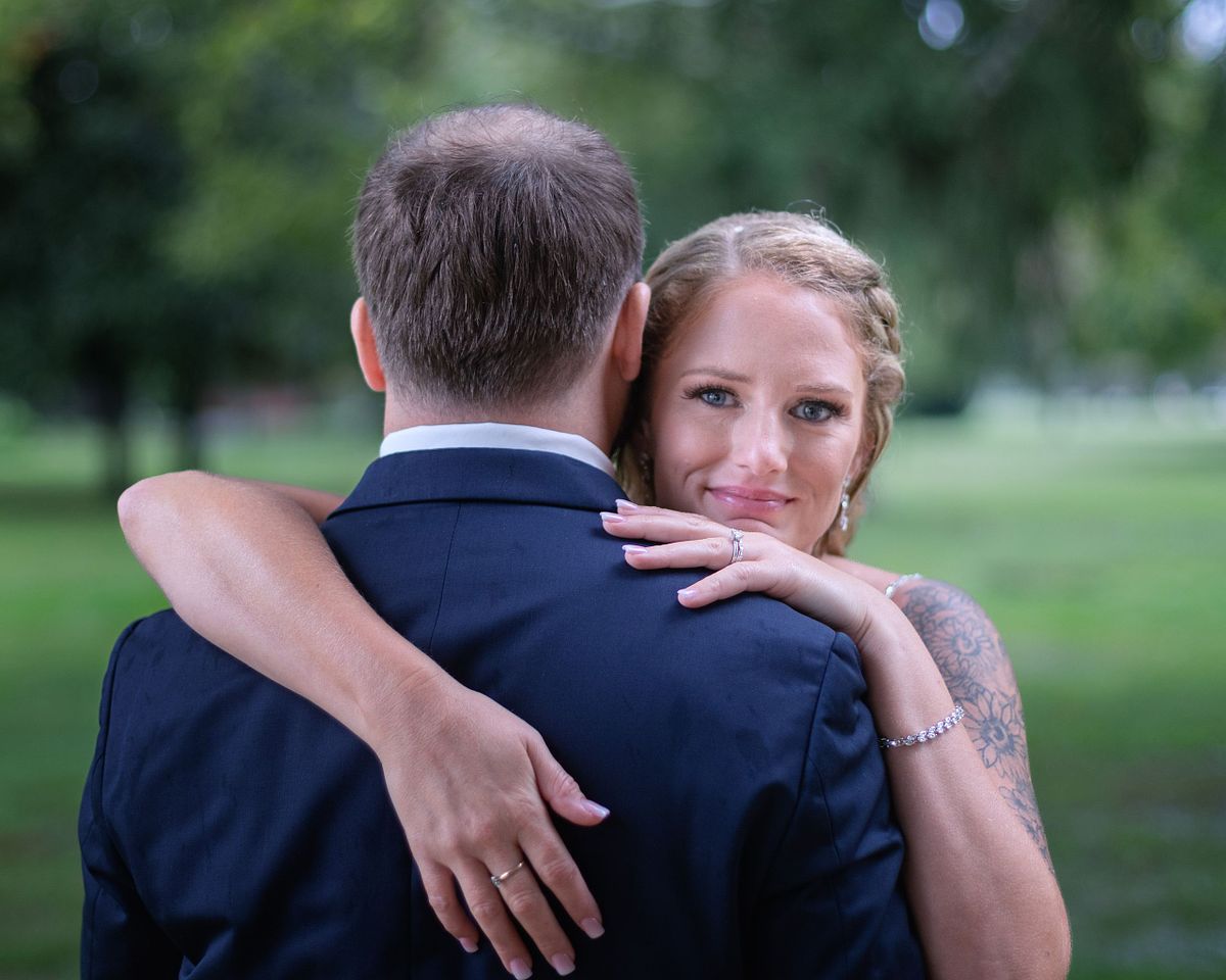 close up of bride looking at the photographer and the groom looking in the opposite direction, the brides chin is resting on the grooms shoulder and she is smilling