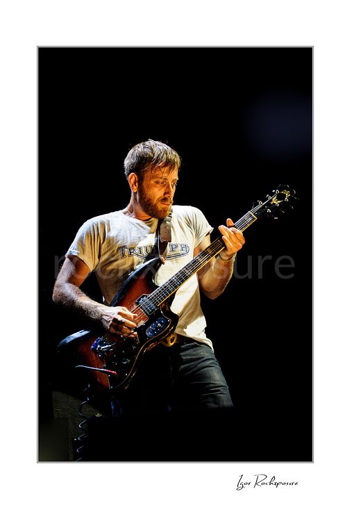 Vertical color image of Dan Auerbach of The Black Keys playing guitar under warm stage lighting in a dark concert scene