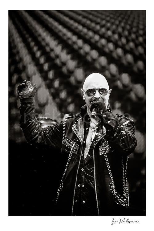Vertical black and white image of Rob Halford of Judas Priest performing live on stage, singing into a microphone while wearing sunglasses and a studded leather coat