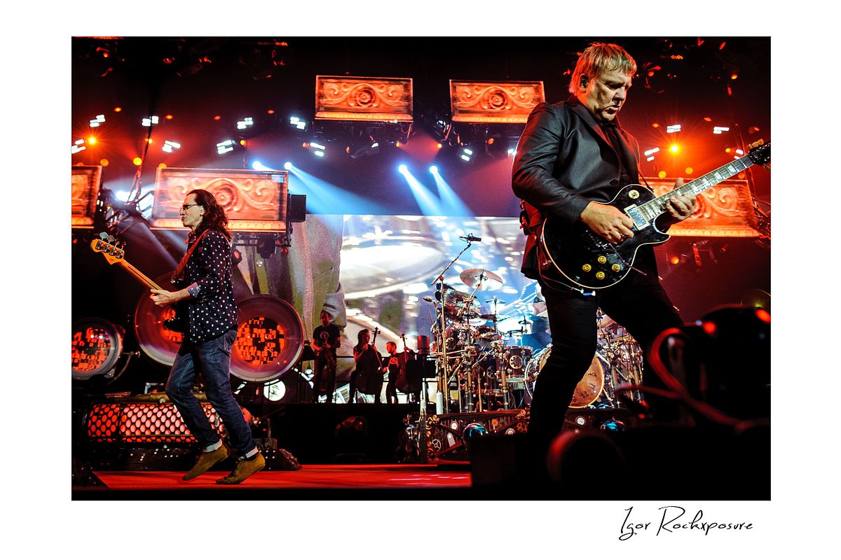 Horizontal concert photography of Rush performing on stage with Alex Lifeson and Geddy Lee under red and blue stage lighting