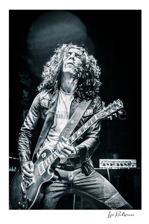 Vertical black and white image of Brent Woods playing guitar on stage with curly hair and dramatic overhead light