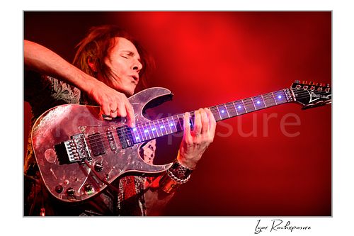 Horizontal color image of Steve Vai playing guitar in close-up under vivid red stage lighting with illuminated fret markers