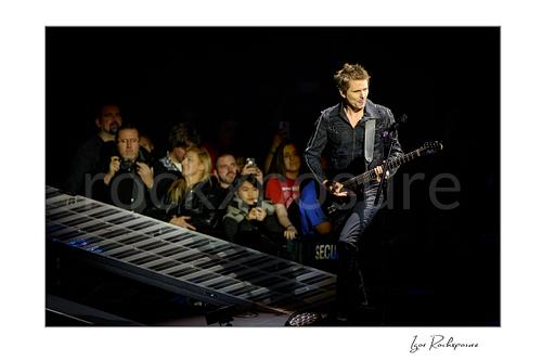 Horizontal color image of Matt Bellamy of Muse playing guitar beside a piano-key stage ramp before the arena crowd