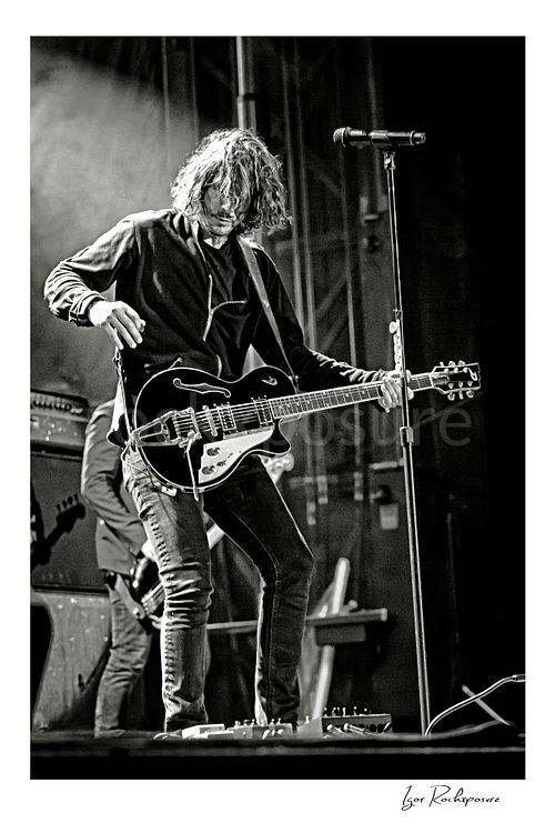 Vertical black and white image of Chris Cornell performing live on stage, playing electric guitar beside a microphone stand under concert lighting