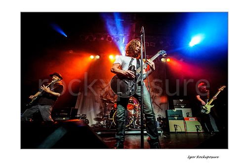 Horizontal color image of Kim Thayil, Chris Cornell, and Ben Shepherd of Soundgarden performing live on stage, with Thayil at left, Cornell at center singing and playing guitar, and Shepherd at right under vivid red, orange, and blue concert lighting