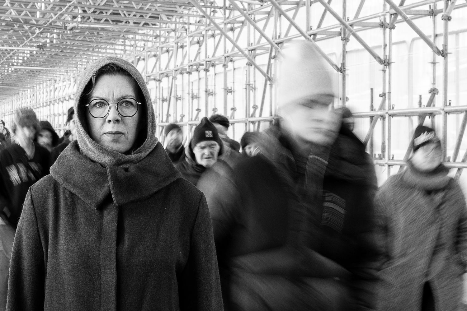Conceptual black and white portrait of a woman in a hooded coat standing still among a moving crowd, photographed in Stockholm by Mats Karlsson.