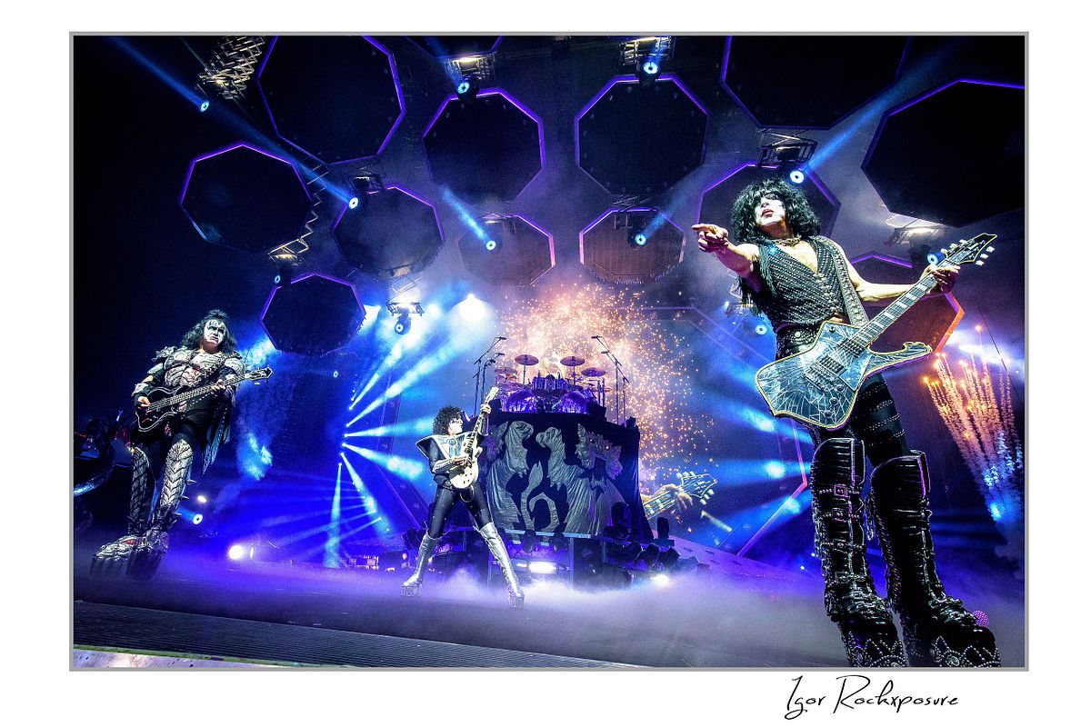 Horizontal color photograph of Gene Simmons, Paul Stanley, and Tommy Thayer performing onstage in full makeup and costumes under intense blue and purple lighting with pyrotechnic smoke and hexagonal overhead lights