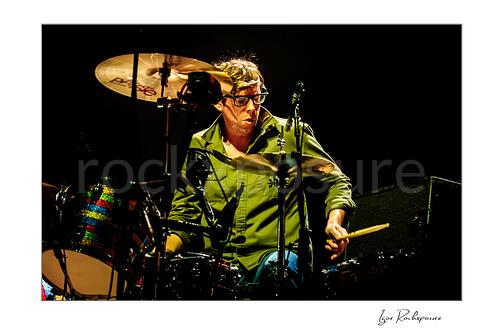 Horizontal color image of Patrick Carney of The Black Keys playing drums under warm stage lighting in a dark concert scene