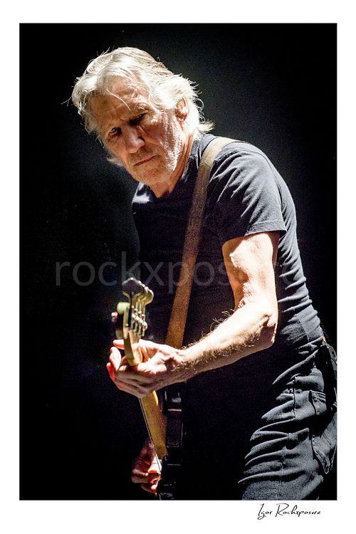 Vertical color image of Roger Waters performing live on stage in a close concert portrait, playing bass under dramatic spotlight against a dark background