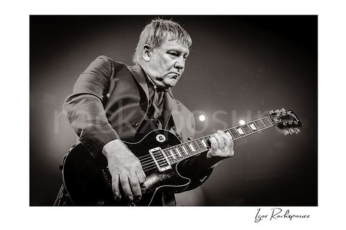 Horizontal black and white image of Alex Lifeson performing with electric guitar during a Rush concert under dramatic stage light