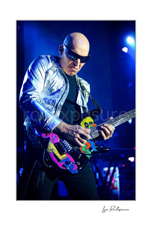 Vertical color image of Joe Satriani performing live in silver jacket and sunglasses with vividly painted guitar under blue stage lights