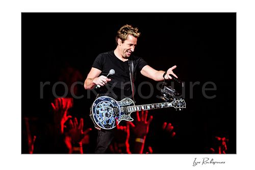Horizontal color image of Chad Kroeger of Nickelback holding a microphone and guitar while reaching toward the crowd under red concert light