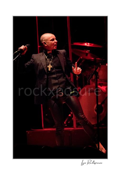 Vertical color image of Sinéad O’Connor holding a microphone on stage with the drum kit behind her under red lighting