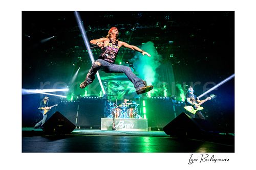 Horizontal color image of Poison performing live on stage with C.C. DeVille, Bret Michaels, Rikki Rockett and Bobby Dall, with Bret Michaels leaping midair under vivid green stage light and smoke