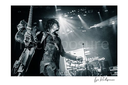 Horizontal black and white image of Tom Keifer on stage holding his guitar upright in a dramatic live performance moment, with bright concert beams and the drum kit visible behind him