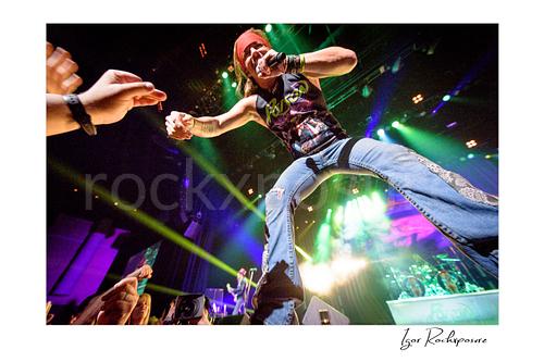 Horizontal color image of Bret Michaels performing live during a Poison concert, leaning toward the crowd with microphone in hand under vivid stage lights