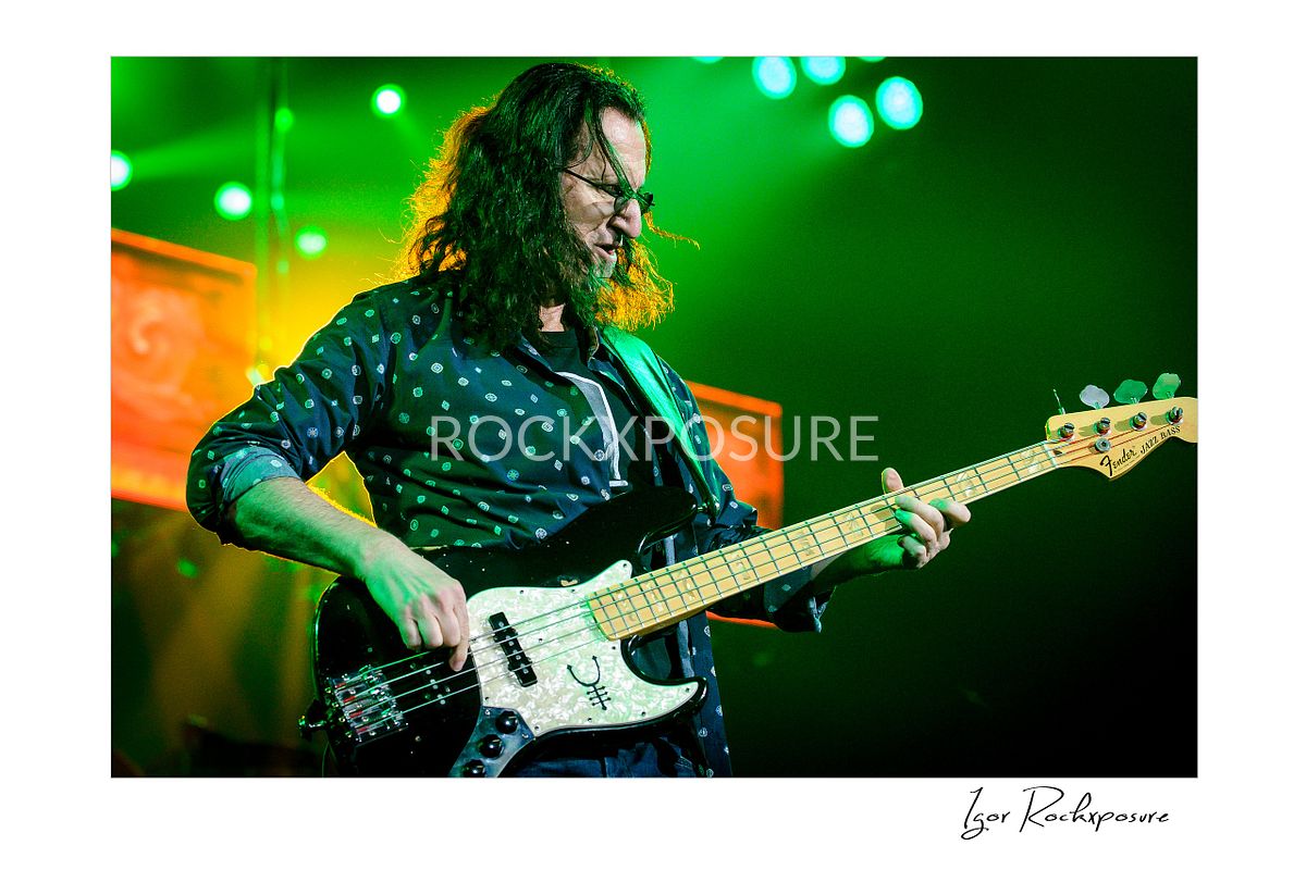 Horizontal concert photography of Geddy Lee playing bass guitar under green stage lighting