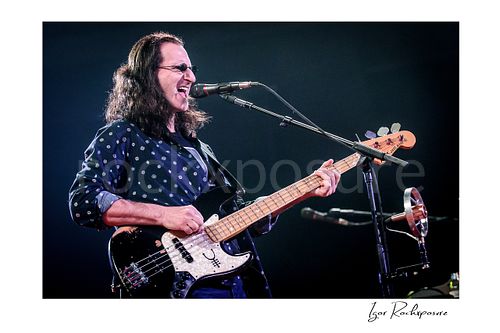 Horizontal color image of Geddy Lee singing into a microphone while playing bass guitar during a Rush concert under bright stage light