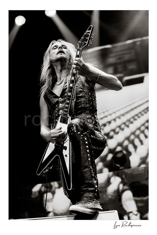 Vertical black and white image of Richie Faulkner of Judas Priest performing live on stage, playing flying V guitar in a dramatic low-angle shot under bright concert lighting