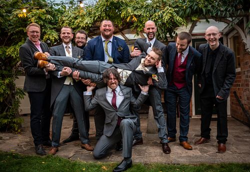 The groom being held sideways by his groomsmen in a playful and fun wedding photo, captured by Weddings By Jermaine.