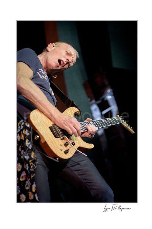 Vertical color image of Phil Collen of Def Leppard performing live on stage, playing electric guitar in a dramatic low-angle shot under concert lighting