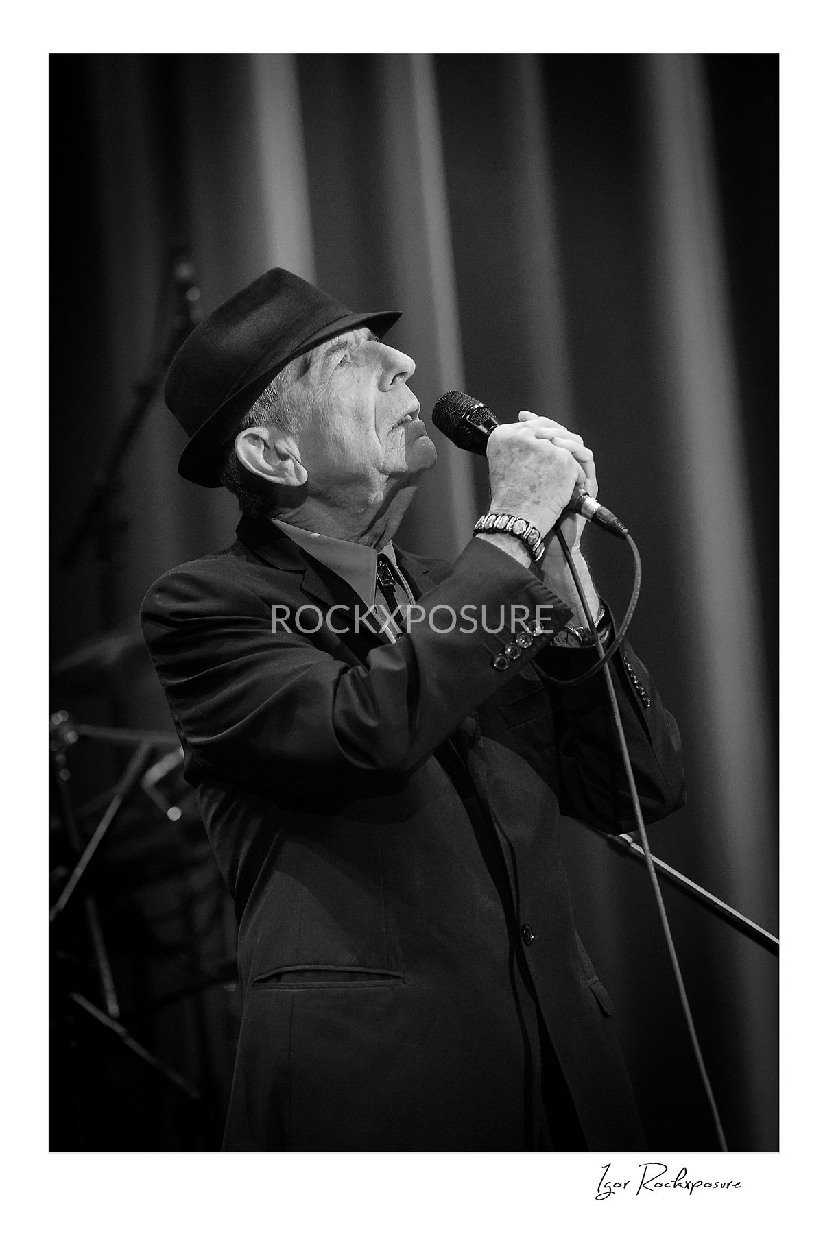 Vertical concert photography of Leonard Cohen singing into a microphone in black and white photography under stage lighting