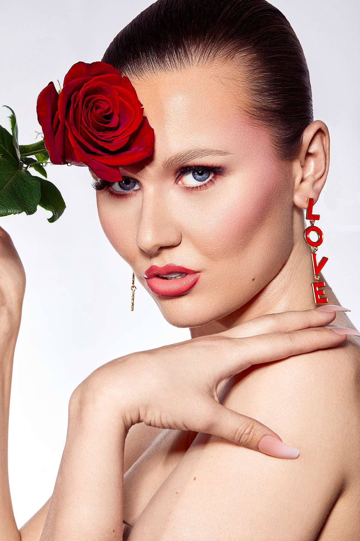 Portrait of a woman with a red rose covering one eye and 'LOVE' drop earrings.