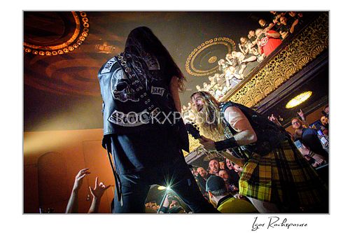 Dario Lorina seen from behind wearing a denim vest, standing on stage next to Zakk Wylde who is leaning forward playing guitar.