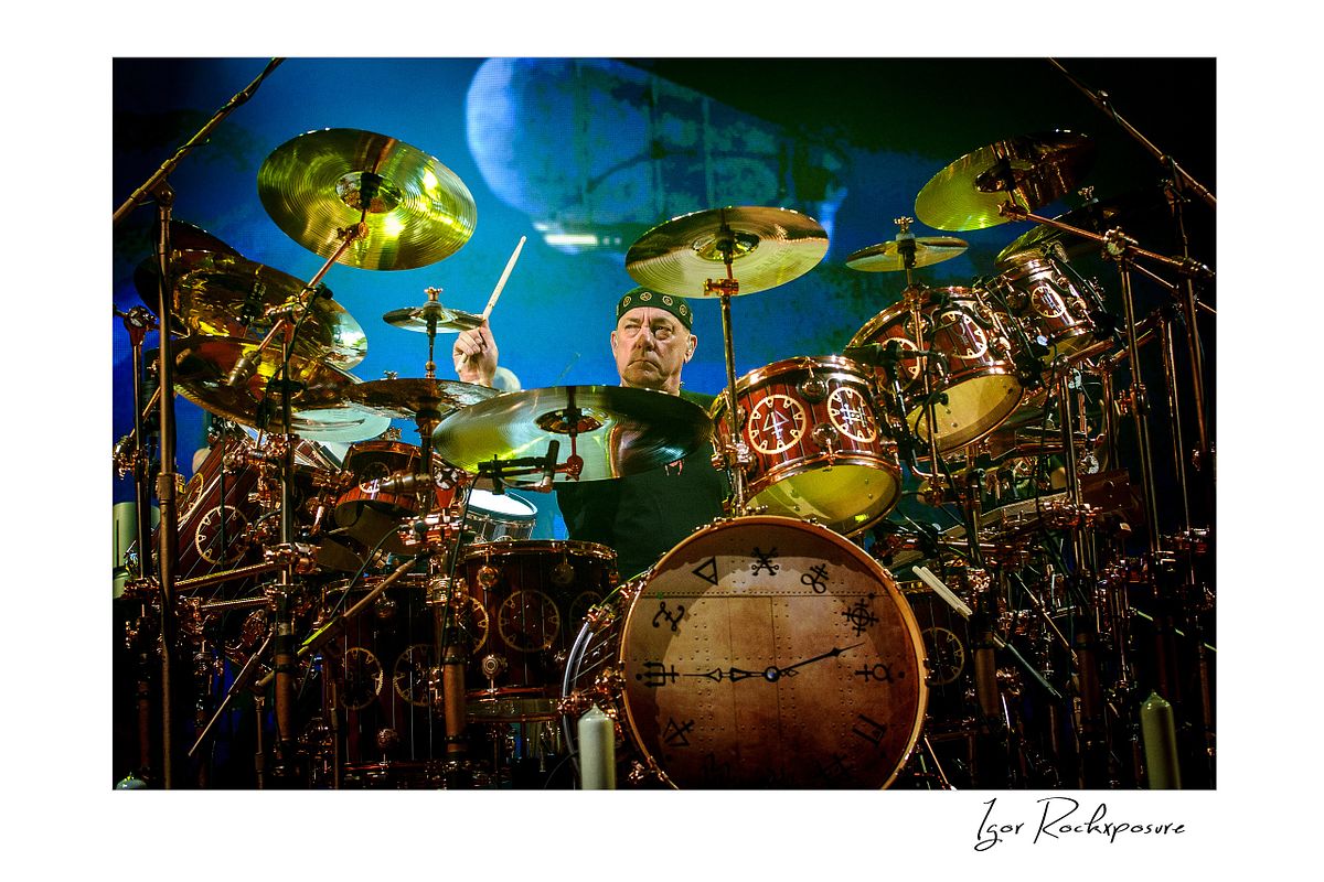 Horizontal concert photography of Neil Peart performing behind a large drum kit surrounded by cymbals under colorful stage lighting