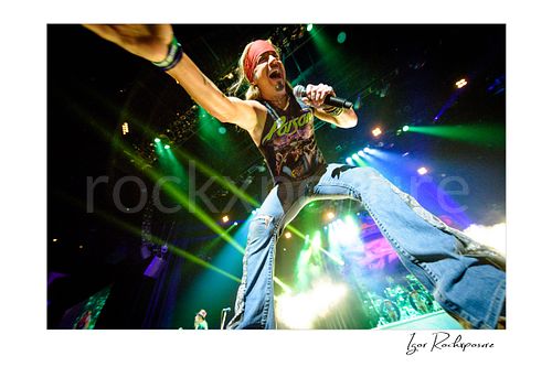 Horizontal color image of Bret Michaels performing live during a Poison concert, reaching toward the crowd with microphone in hand under vivid green stage lights