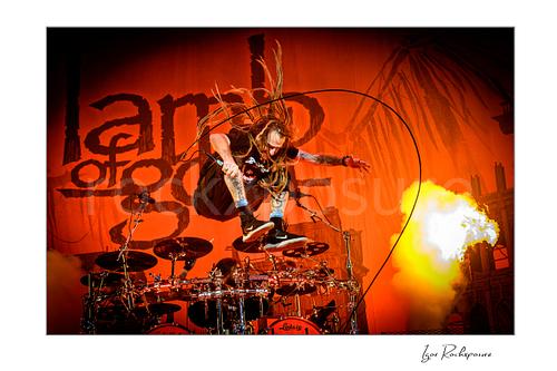 Horizontal color image of Randy Blythe of Lamb of God leaping above the drum kit with flying dreadlocks, microphone cable looping through the air, and a burst of stage fire