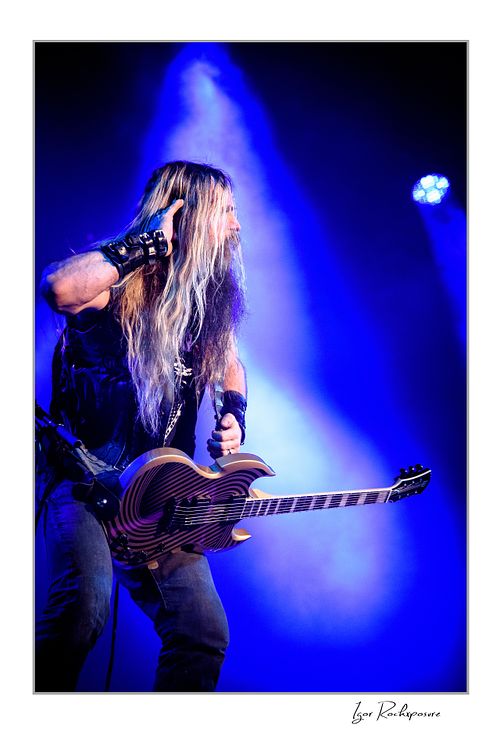Vertical shot of Zakk Wylde playing guitar on stage, wearing a denim vest, looking to the side under an intense blue spotlight and atmospheric stage fog.
