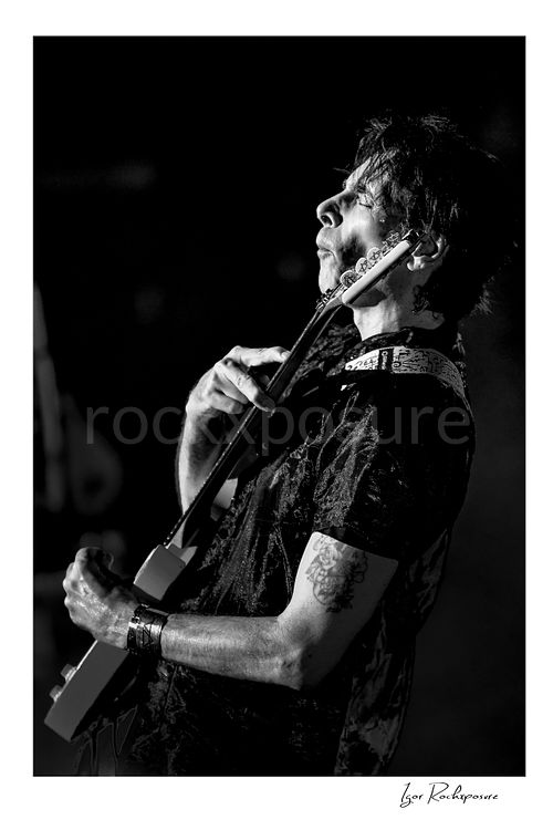 Vertical black and white image of Steve Vai playing guitar in close-up with head tilted back and an expressive stage pose
