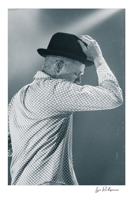 Vertical black and white image of Gord Downie of The Tragically Hip in a quiet live performance moment, touching the brim of his hat under soft stage light