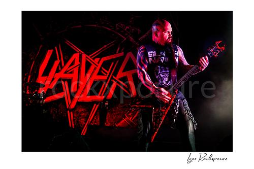 Horizontal color image of Kerry King of Slayer playing guitar onstage under intense red and blue concert lighting with the Slayer logo behind him