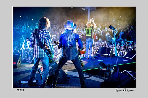 Horizontal black and white image of Bret Michaels leaping in midair with Bobby Dall and C.C. DeVille performing during a Poison concert in a dramatic backlit stage scene